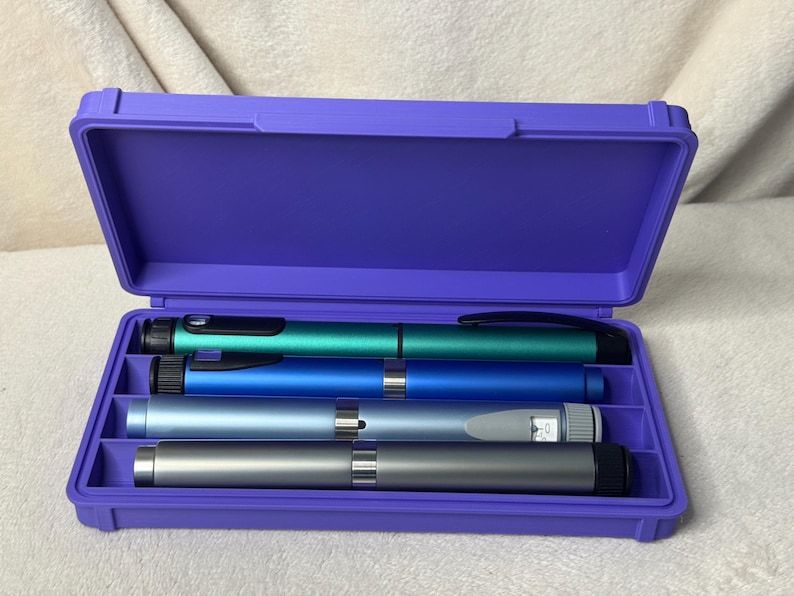 May include: A purple plastic case with an open lid, containing four insulin pens in shades of teal, blue, and silver. The pens are arranged neatly within the case, which is set against a neutral background.