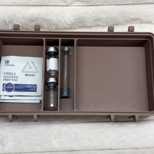 May include: A rectangular brown plastic storage case with three internal compartments. The left section holds two white sterile alcohol prep pad packets. The center section contains two small glass vials with silver caps and one thin glass tube.