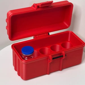 May include: A red plastic case with a hinged lid, designed to hold small vials or bottles. The case has four compartments and a small blue lidded vial is visible inside.