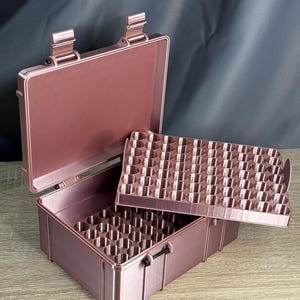 May include: A rose gold, open, rectangular case with a removable tray. The case has a hinged lid and a black latch. The interior of the case and tray have multiple cylindrical compartments.