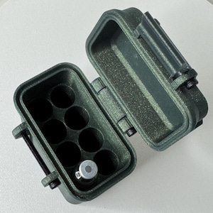 May include: An open, olive-green, rectangular case with a hinged lid. The interior has nine circular compartments, one of which contains a small, silver-colored object. The case is made of a textured material and has a secure latch.