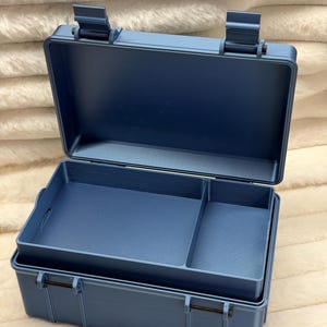 May include: A matte blue rectangular storage case with a hinged lid and two secure latches. The interior features a removable tray with two distinct compartments, providing organized storage space within the sturdy, structured container design.