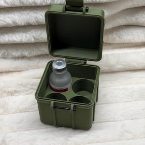 May include: An olive green, square, hinged storage case with a latch mechanism. The interior features four circular cutouts designed to hold small vials. One small, translucent glass vial with a silver cap is placed in one of the circular slots.