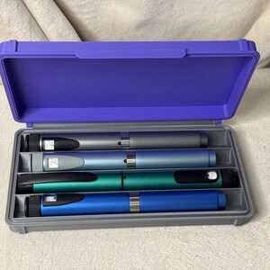 May include: A purple plastic case with a gray interior holds four insulin pens in silver, blue, teal, and blue. The case is open, revealing the pens, which have black caps and digital displays.