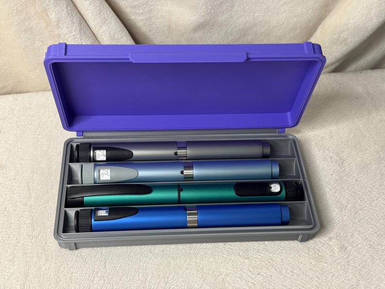 May include: A purple plastic case with a gray interior holds four insulin pens in silver, blue, teal, and blue. The case is open, revealing the pens, which have black caps and digital displays.