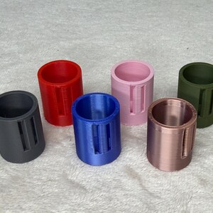 May include: A collection of six cylindrical objects in various colors: dark gray, red, blue, pink, copper, and olive green. Each cylinder has a rectangular cutout. The objects are arranged on a white, textured surface.