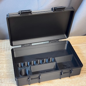 May include: Open black plastic case with a hinged lid and secure latches. The interior features compartments and cylindrical holders. The case is designed for organizing and storing small items, tools, or equipment. The case is sitting on a wooden surface.
