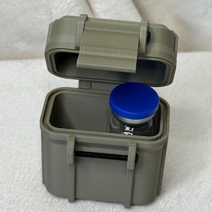 May include: An open, olive-green, small, rectangular case with a hinged lid and a black latch. Inside the case is a small glass vial with a blue cap. The case is sitting on a white, textured surface.