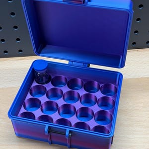 May include: A blue and purple storage case with an open lid. The interior has multiple circular compartments. A small bottle sits in one of the compartments. The case has a handle and a latch. The case is on a wooden surface.