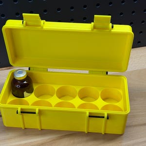 May include: Bright yellow plastic case with an open lid, featuring a brown glass bottle with a silver cap. The interior has circular compartments. The case is designed with two latches and two hinges.