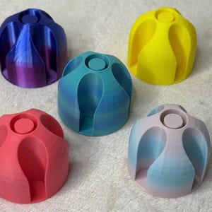 May include: Five colorful, 3D-printed bottle stoppers. The stoppers are in shades of blue, pink, yellow, and red. Each stopper has a rounded shape with a circular top and curved sides. The objects are sitting on a white, textured surface.