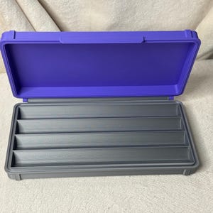 May include: An open, two-tone storage case with a purple lid and a gray base. The interior of the base features multiple gray compartments. The case is designed to hold and organize small items.