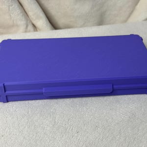 May include: A closed, rectangular, purple case with a textured surface. The case has a handle and reinforced corners. The case is sitting on a light-colored, textured surface.