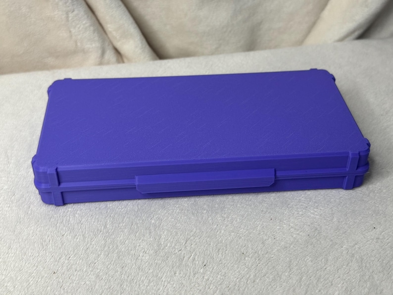 May include: A closed, rectangular, purple case with a textured surface. The case has a handle and reinforced corners. The case is sitting on a light-colored, textured surface.