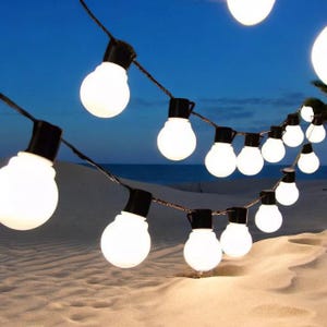 May include: A string of white globe lights with black cords hanging from a sandy beach at dusk. The lights are strung in a curved line and are illuminated against the blue sky.