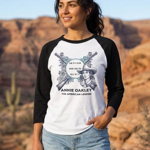 May include: A white baseball t-shirt with black three-quarter sleeves featuring a graphic of Annie Oakley in a cowboy hat, two crossed rifles, and roses. The text reads: AIM AT A HIGH MARK AND YOU WILL HIT IT, ANNIE OAKLEY, THE AMERICAN LEGEND.