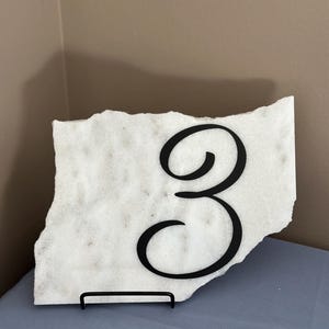 Peut inclure: Numéro de table en marbre blanc avec un support en métal noir et un numéro "3" en métal noir en écriture cursive.