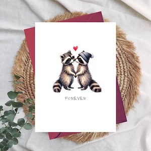 May include: A white greeting card featuring two raccoon illustrations, touching noses and holding hands, with a red heart above them. One raccoon wears a blue and white striped hat. The word "FOREVER" is printed below the raccoons.