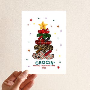 Puede incluir: Tarjeta de Navidad con un diseño de árbol hecho de zapatos Crocs con varios estampados, incluyendo cuadros rojos, verde y estampado de leopardo. Una estrella dorada corona el árbol, con la frase "CROCIN' AROUND THE CHSRISTMAS TREE" debajo. Estrellas coloridas rodean el árbol.