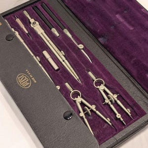 May include: A vintage drafting set in a black leather case with a purple velvet interior. The set includes several silver-coloured compasses, dividers, and other drawing tools. The case is marked "POST MADE IN U.S.A."
