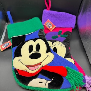 May include: Two Christmas stockings featuring Mickey and Minnie Mouse. The Mickey Mouse stocking has a green cuff, blue scarf, and red accents. The Minnie Mouse stocking has a purple cuff and a holly design. Both stockings have a loop for hanging.