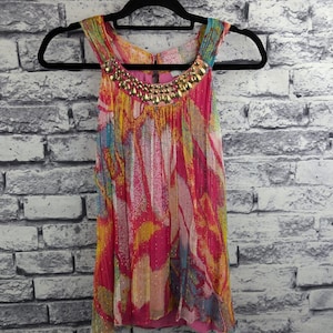 以下が含まれることがあります： ピンク、黄色、青の色合いの鮮やかな抽象模様のノースリーブトップス。ネックラインは金色のビーズで飾られています。白いレンガの壁を背景に、黒いハンガーに吊るされたトップスです。