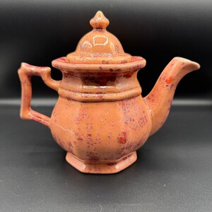 Puede incluir: Una pequeña tetera de cerámica con tapa y asa. La tetera es de color naranja moteado con detalles en rojo. El pico y el asa son curvos. La tapa tiene un pequeño adorno en la parte superior. La tetera tiene una base hexagonal.
