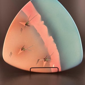 May include: A triangular ceramic plate with a blue and pink glaze. The plate features a design of three stylised dragonflies in shades of green and brown.