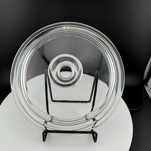 Könnte beinhalten: Ein durchsichtiger Glasdeckel mit einem erhöhten Mittelring. Der Deckel steht auf einem schwarzen Metallständer.