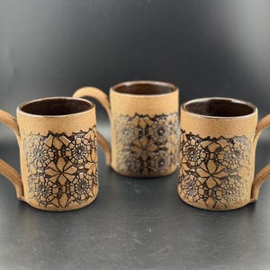 May include: Three cylindrical ceramic mugs with a textured, light brown exterior featuring a dark brown, embossed geometric floral pattern. Each mug has a curved handle and a smooth, dark brown interior finish, arranged in a group of three.