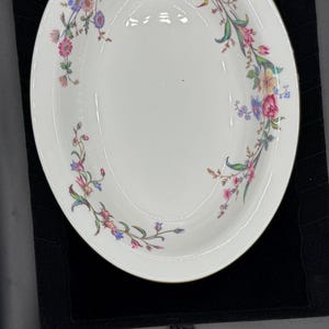 May include: White, oval-shaped serving bowl with a floral design. The bowl features a gold rim and is decorated with pink, blue, and yellow flowers and green leaves. The floral pattern is arranged around the rim.
