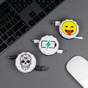 Peut inclure: Trois câbles de chargement USB rétractables blancs et noirs avec différents motifs : un visage souriant jaune, une icône de batterie et une tête de mort. Les câbles sont sur une surface grise près d'un clavier et d'une souris.