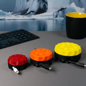 Op de afbeelding: Drie kabelorganisatoren in verschillende kleuren: rood, oranje en geel. Elke organisator heeft een ronde basis met een gestructureerd oppervlak en een kabelgleuf. De organisatoren bevinden zich op een grijs oppervlak.