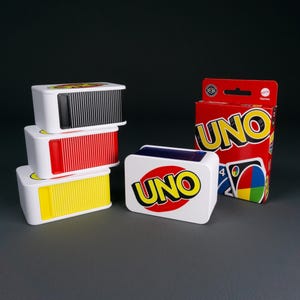 Könnte beinhalten: Ein Stapel aus drei rechteckigen Kartenhaltern aus weißem Kunststoff mit schwarzen, roten und gelben gerippten Schiebedeckeln sowie ein einzelner weißer Kartenhalter mit dem UNO-Logo und eine rote UNO-Spielkartenschachtel mit Markenname und Grafik.
