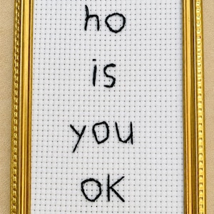 May include: A rectangular gold-toned frame containing white Aida fabric with a grid pattern. Black thread is used to hand-stitch the text "ho is you ok" in a simple, lowercase font centered within the frame, creating a wall-mounted piece of art.