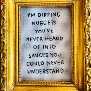 Puede incluir: Una obra de arte en punto de cruz enmarcada con un marco dorado ornamentado. El texto dice: "I'M DIPPING NUGGETS YOU'VE NEVER HEARD OF INTO SAUCES YOU COULD NEVER UNDERSTAND" en hilo negro sobre fondo blanco.