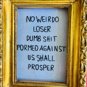 May include: A framed cross-stitch piece with a gold ornate frame. The cross-stitch is light blue with black text that reads "NO WEIRDO LOSER DUMB SHIT FORMED AGAINST US SHALL PROSPER."