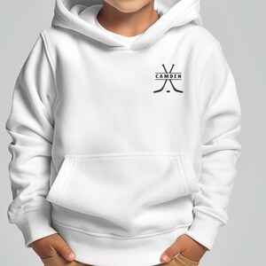 May include: A white hooded sweatshirt featuring a black graphic on the upper left chest. The graphic consists of two crossed hockey sticks with the word CAMDEN printed in black capital letters inside a horizontal rectangular box between them.