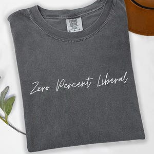 May include: A dark gray t-shirt with the text "Zero Percent Liberal" in white script. The shirt has a crew neck and a tag that reads "Comfort Colors". A brown hat is partially visible in the background.