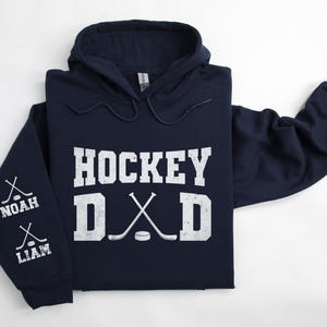Peut inclure: Un sweat &agrave; capuche bleu marine avec les mots "HOCKEY DAD" en lettres blanches. Les manches ont des crosses de hockey et les noms "NOAH" et "LIAM". Le sweat &agrave; capuche a une capuche &agrave; cordon et une poche kangourou.