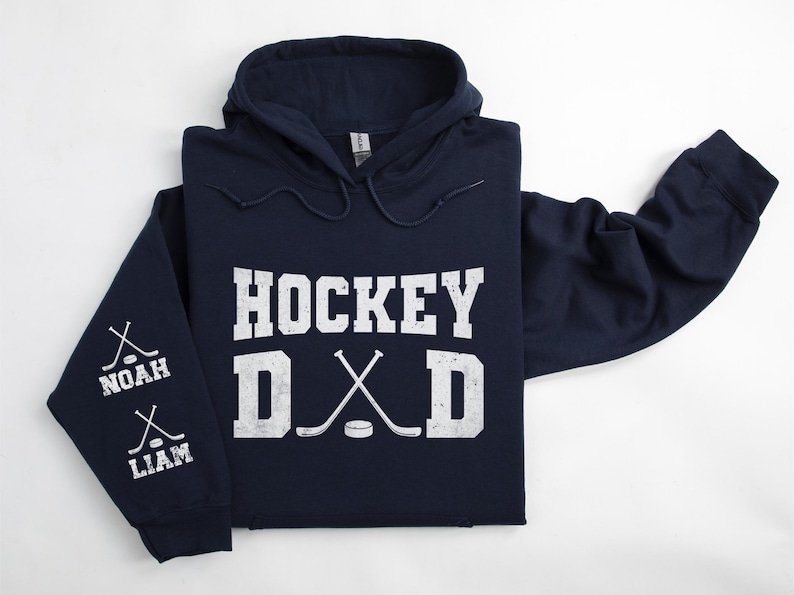 Peut inclure: Un sweat &agrave; capuche bleu marine avec les mots "HOCKEY DAD" en lettres blanches. Les manches ont des crosses de hockey et les noms "NOAH" et "LIAM". Le sweat &agrave; capuche a une capuche &agrave; cordon et une poche kangourou.