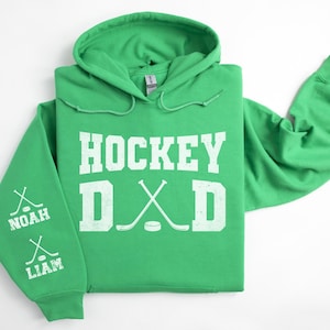 Peut inclure: Un sweat &agrave; capuche vert avec l'inscription blanche "HOCKEY DAD". Le design comprend des crosses et une rondelle de hockey. La manche gauche affiche les noms "NOAH" et "LIAM" avec des graphiques de crosses.
