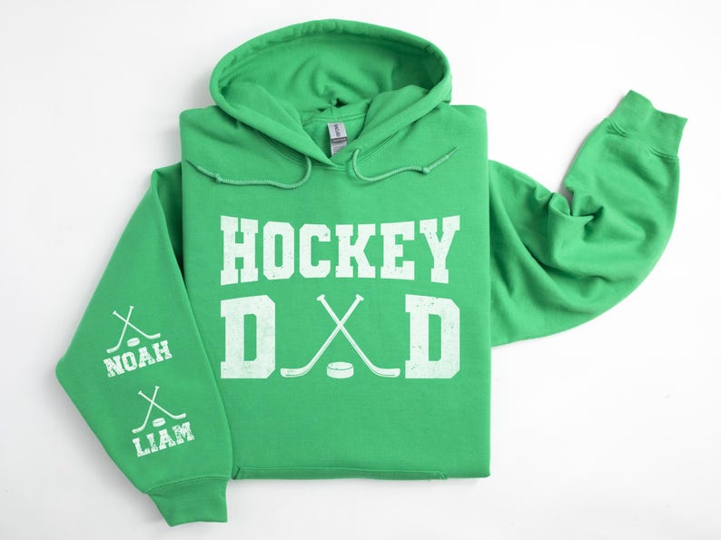 Peut inclure: Un sweat &agrave; capuche vert avec l'inscription blanche "HOCKEY DAD". Le design comprend des crosses et une rondelle de hockey. La manche gauche affiche les noms "NOAH" et "LIAM" avec des graphiques de crosses.