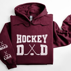 Peut inclure: Un sweat &agrave; capuche bordeaux avec l'inscription blanche "HOCKEY DAD". Les manches affichent des crosses de hockey crois&eacute;es et les noms "NOAH" et "LIAM". Le sweat &agrave; capuche a une capuche avec des cordons.