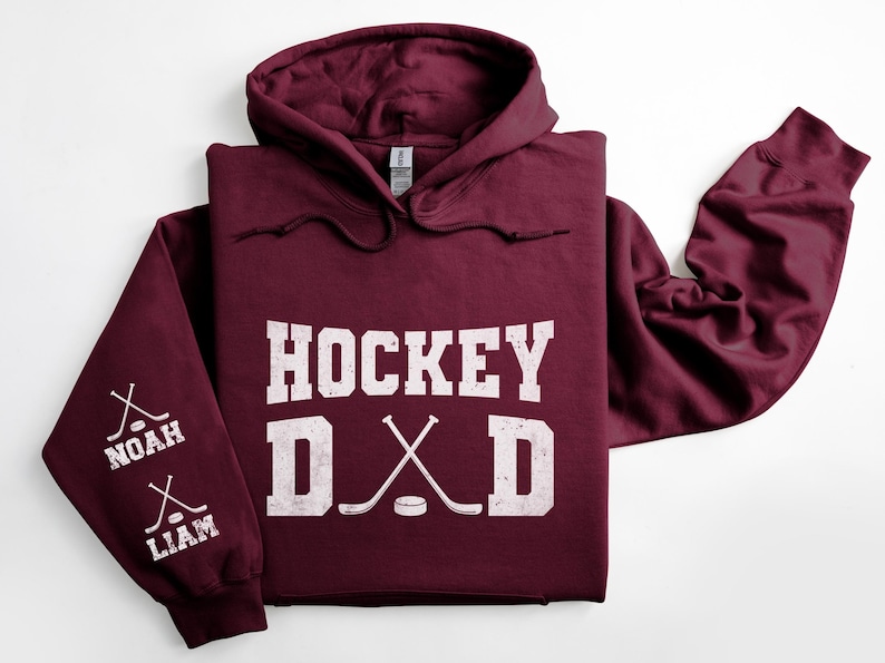Peut inclure: Un sweat &agrave; capuche bordeaux avec l'inscription blanche "HOCKEY DAD". Les manches affichent des crosses de hockey crois&eacute;es et les noms "NOAH" et "LIAM". Le sweat &agrave; capuche a une capuche avec des cordons.