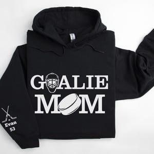 May include: Black hoodie with white text and graphics. The front displays "GOALIE MOM" with a goalie mask and hockey puck graphic. The left sleeve has crossed hockey sticks, the name "Evan" and the number "53".