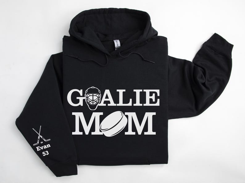 May include: Black hoodie with white text and graphics. The front displays "GOALIE MOM" with a goalie mask and hockey puck graphic. The left sleeve has crossed hockey sticks, the name "Evan" and the number "53".