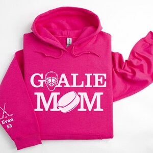 May include: A bright pink hoodie with the words "GOALIE MOM" in white, along with a hockey mask and puck graphic. The left sleeve has crossed hockey sticks and the name "Evan 53".