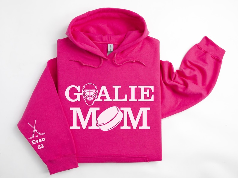 May include: A bright pink hoodie with the words "GOALIE MOM" in white, along with a hockey mask and puck graphic. The left sleeve has crossed hockey sticks and the name "Evan 53".
