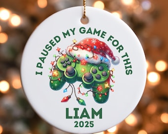 May include: White round Christmas ornament with a green video game controller design. The controller is decorated with Christmas lights and a Santa hat. Text reads "I PAUSED MY GAME FOR THIS" and "LIAM 2025".